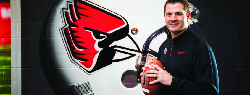 Ball State football coach Mike Neu holds a football while standing in front of a mural of a team football helmet.