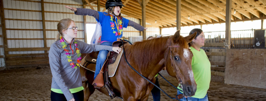 Two adults help a young girl as she rides a horse in a barn.