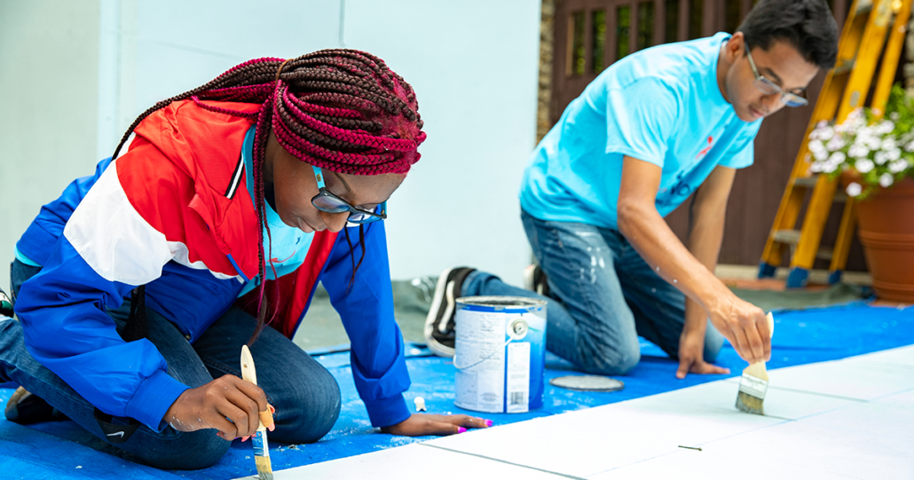 A teenage girl and boy paint some wood from a crouched position