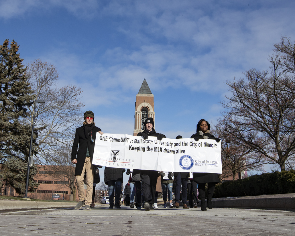 Several people in coats and hats march peacefully down a road while holding a banner that says "One Community: Ball State University and the City of Muncie Keeping the MLK dream alive"