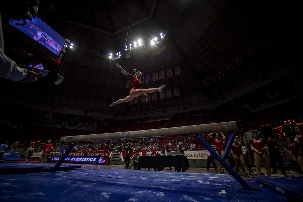 A gymnast does a leap split above a balance beam, surrounded by her teammates and a crowd