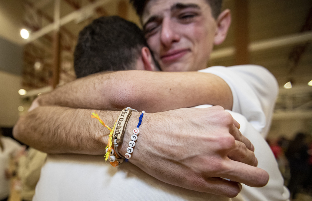 Two students hug in a gymnasium full of people, tears and emotion evident on one student's face