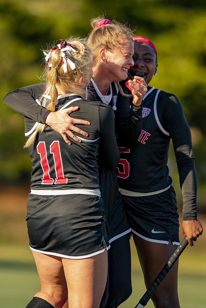 Three field hockey players embrace on the field, their faces full of emotion